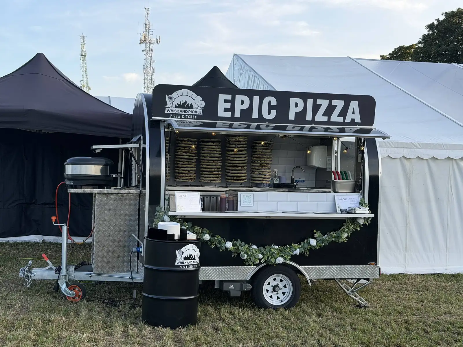 The Whisk And Pickle mobile pizza kitchen set up on grass beside a marquee, stacked pizza bases behind the serving hatch and an EPIC PIZZA sign above it.