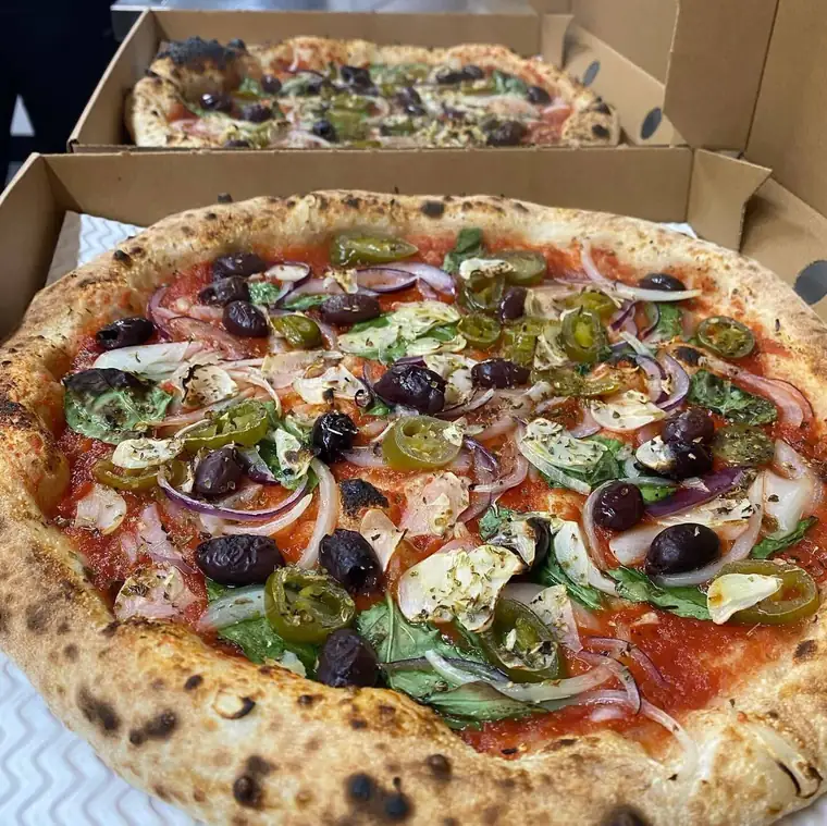 A pizza loaded with olives, jalapenos, red onion and basil, a second box stacked behind it.